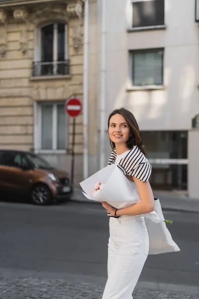 Cheerful young woman in stylish outfit holding bouquet wrapped in paper on street in paris — Stock Photo