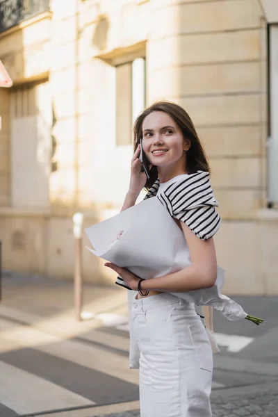 Felice giovane donna in abito elegante parlando su smartphone e tenendo in mano bouquet avvolto in carta su strada a Parigi — Foto stock