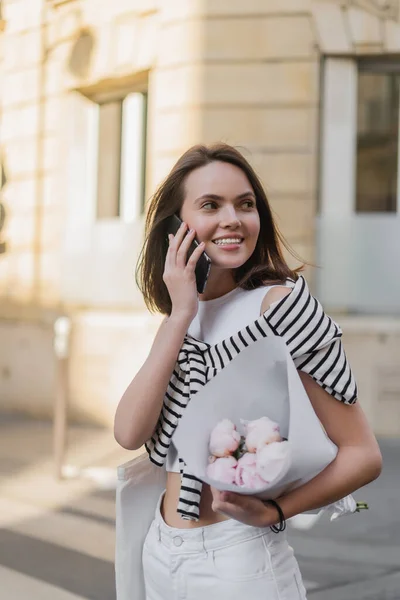 Mujer alegre en traje elegante hablando en el teléfono inteligente y la celebración de ramo con peonías en la calle en París - foto de stock