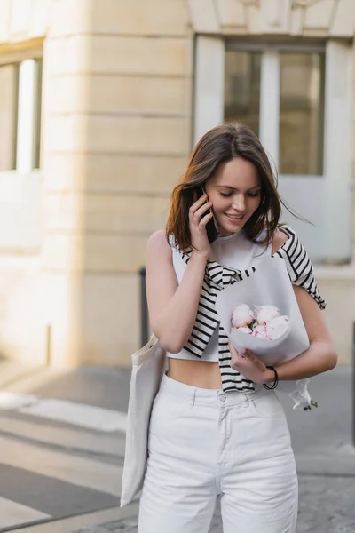 Pleased woman in stylish outfit talking on smartphone and holding bouquet with peonies on street in paris — Stock Photo