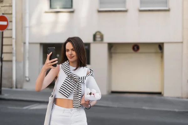 Весела жінка в стильному вбранні дивиться на смартфон і тримає букет з півоніями на вулиці в Парижі — стокове фото
