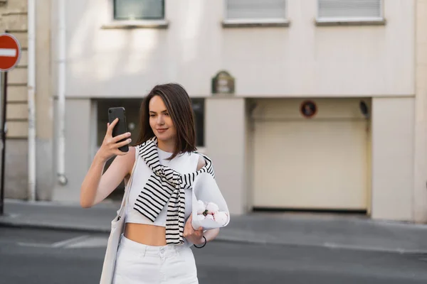 Mujer alegre en traje elegante mirando el teléfono inteligente y la celebración de ramo con peonías en la calle en París - foto de stock