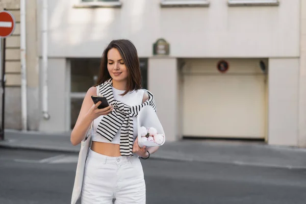 Веселая женщина в стильном наряде, используя смартфон и держа букет с пионами на улице в Париже — стоковое фото