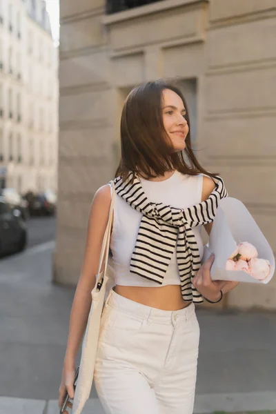 Задоволена молода жінка в стильному вбранні тримає букет квітучих півоній, загорнутий в папір на вулиці в Парижі — стокове фото