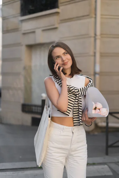 Femme heureuse en tenue tendance parlant sur téléphone portable et tenant un bouquet avec des pivoines dans la rue à Paris — Photo de stock