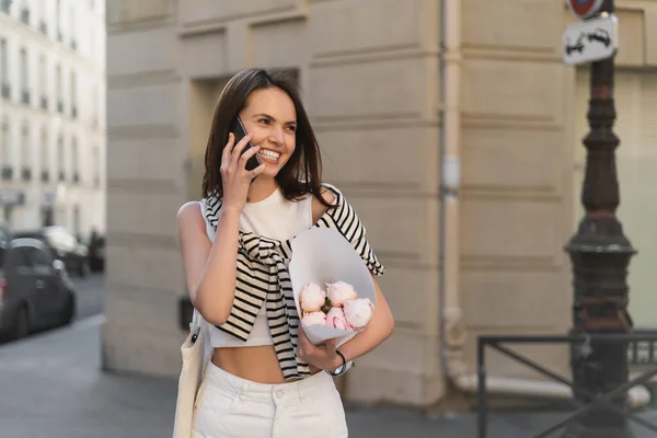 Positive woman in trendy outfit talking on mobile phone and holding bouquet with peonies on street in paris — Stock Photo