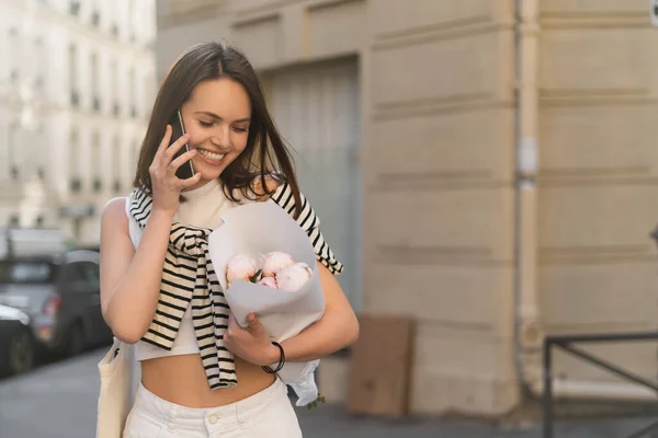 Femme heureuse en tenue tendance parlant sur téléphone portable et tenant un bouquet avec des pivoines dans la rue urbaine à Paris — Photo de stock
