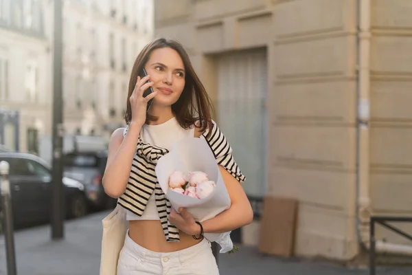 Lächelnde Frau, die mit dem Handy telefoniert und einen Strauß mit Pfingstrosen auf einer städtischen Straße in Paris hält — Stockfoto