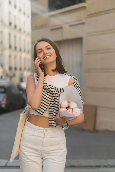 Mulher satisfeita falando no telefone celular e segurando buquê com peônias na rua urbana em paris — Fotografia de Stock