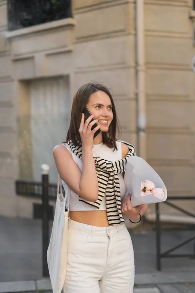 Femme heureuse en tenue tendance parlant sur téléphone portable et tenant des pivoines dans la rue urbaine à Paris — Photo de stock