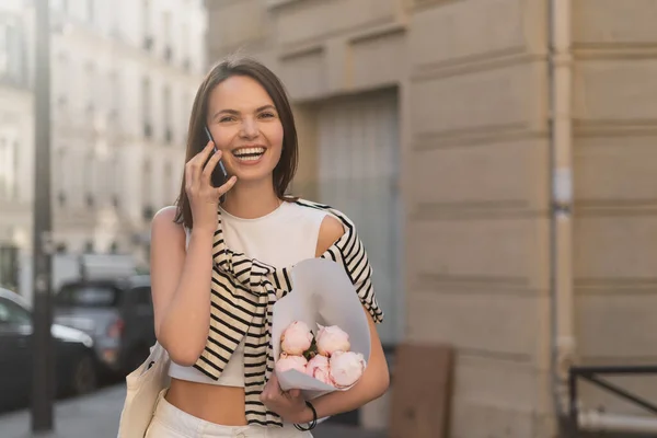 Happy woman in trendy outfit talking on mobile phone while holding bouquet and laughing on urban street in paris — Stock Photo