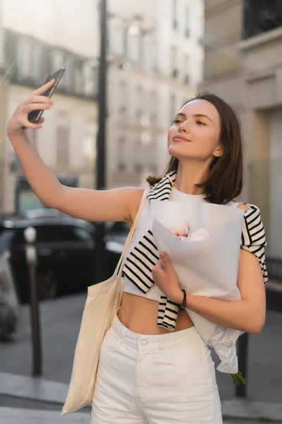 Mujer sonriente en traje de moda tomar selfie en el teléfono inteligente y la celebración de ramo con peonías en la calle en París - foto de stock