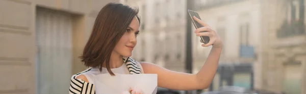 Giovane donna in abito alla moda scattare selfie su smartphone e tenere bouquet con peonie in strada a Parigi, banner — Foto stock