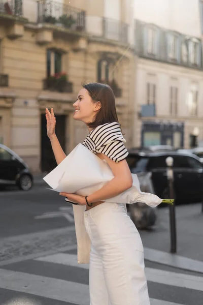 Mulher alegre e elegante segurando buquê de flores envolto em papel enquanto acenando mão na rua em paris — Fotografia de Stock