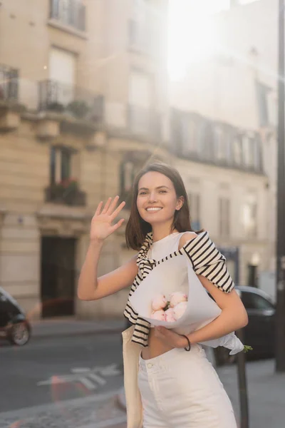 Smiling and stylish woman holding bouquet of flowers wrapped in paper while waving hand on street in paris — Stock Photo