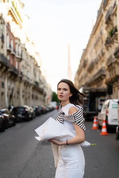 Stylish woman holding bouquet of flowers wrapped in paper while walking on street in paris — Stock Photo