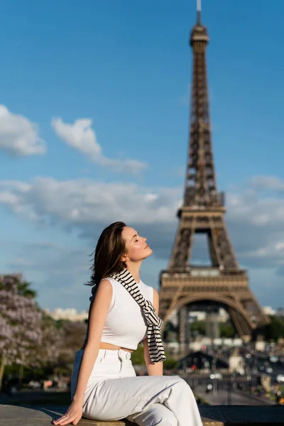 Erfreut junge Frau in stilvollem Outfit sitzt in der Nähe des Eiffelturms in Paris — Stockfoto