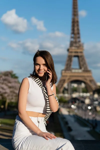 Mujer joven feliz en traje de moda hablando en el teléfono inteligente mientras está sentado cerca de la torre eiffel en París - foto de stock