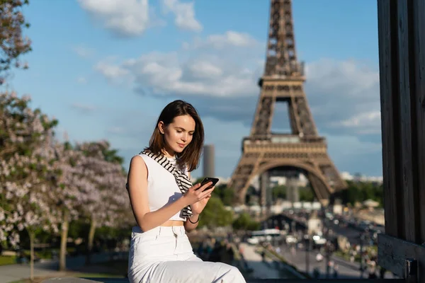 Молодая женщина в стильном наряде, используя смартфон, сидя рядом с Эйфелевой башней в Париже — стоковое фото