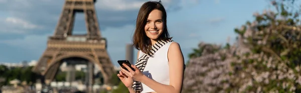 Happy young woman in stylish outfit using smartphone while sitting near eiffel tower in paris, banner — Stock Photo