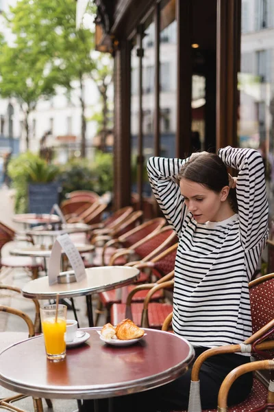 Jeune femme en chemise à manches longues rayée ajustant les cheveux près des boissons et croissant dans un café en plein air à Paris — Photo de stock