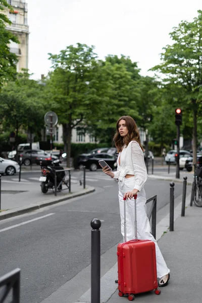 Стильный мужчина с чемоданом и смартфоном стоит у дороги на улице в Париже — стоковое фото
