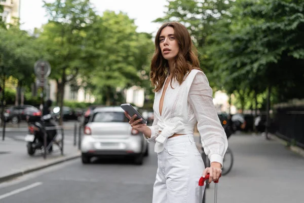 Mujer con estilo con teléfono celular y maleta de pie cerca de la carretera en la calle en París - foto de stock