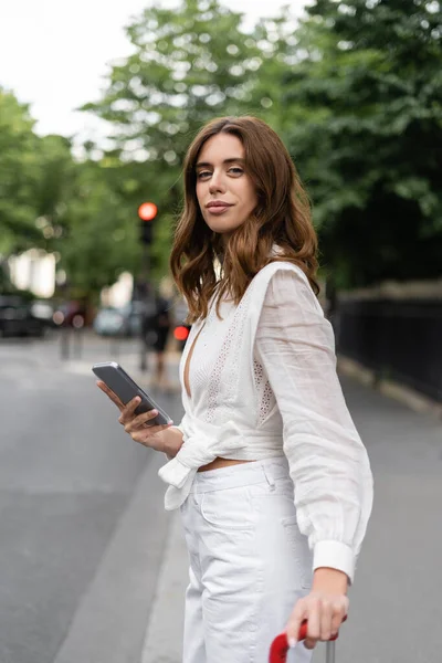 Stylish brunette woman holding smartphone and suitcase on street in Paris — Stock Photo