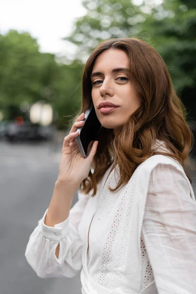 Brunette woman talking on smartphone and looking at camera on street in Paris — Stock Photo