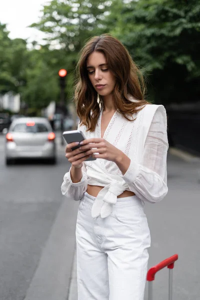 Viajante elegante usando telefone celular perto de mala ao ar livre na França — Fotografia de Stock