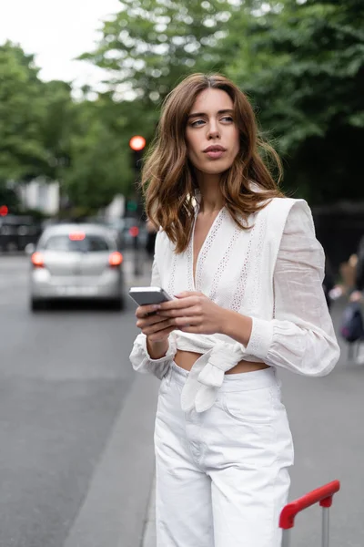 Mujer joven con estilo utilizando el teléfono celular cerca de equipaje en la calle en Francia - foto de stock