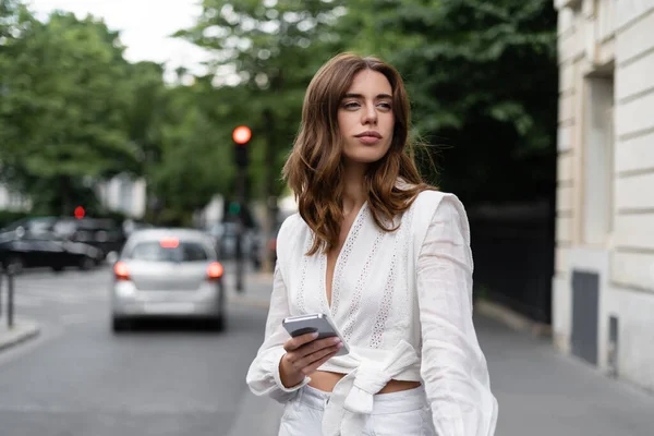 Brunette traveler in blouse holding smartphone near road in Paris — Stock Photo