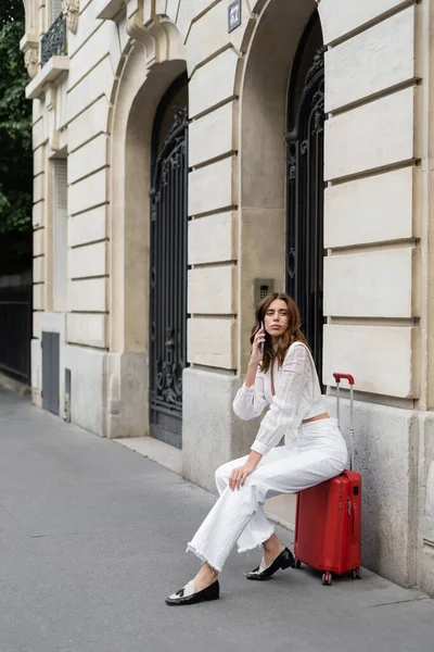 Pretty tourist talking on mobile phone and sitting on suitcase on street in Paris — Stock Photo