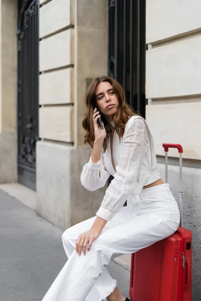 Fashionable traveler talking on smartphone while sitting on baggage on street in Paris — Stock Photo