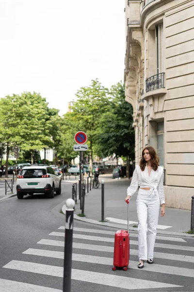 Mujer con estilo en blusa caminando en el paso de peatones cerca de la maleta en la calle en París - foto de stock