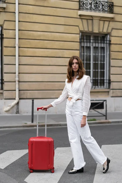 Стильная женщина смотрит в камеру рядом с чемоданом на пешеходном переходе в Париже — стоковое фото