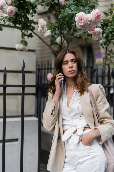 Модная женщина разговаривает по смартфону возле цветущих деревьев на городской улице во Франции — стоковое фото