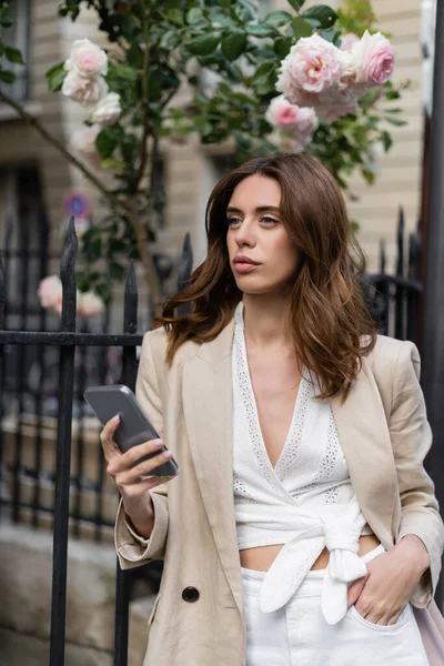 Mujer morena con estilo sosteniendo teléfono inteligente cerca de rosas florecientes en la calle en París - foto de stock