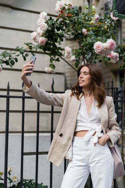 Femme élégante prenant selfie sur smartphone près de roses en fleurs dans la rue à Paris — Photo de stock