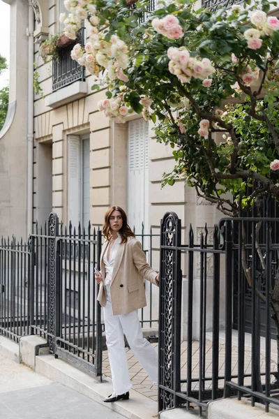 Elegante mujer sosteniendo teléfono inteligente cerca de la valla y el árbol de rosas en flor en la calle en París - foto de stock