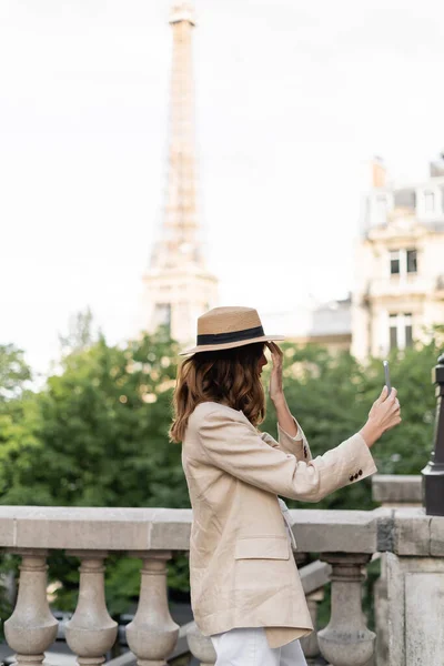 Vista lateral de la mujer con estilo en sombrero de sol tomando selfie en el teléfono inteligente en París - foto de stock