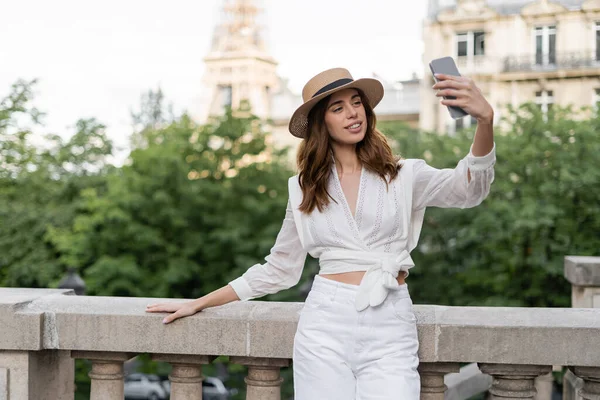 Femme joyeuse en chapeau de soleil prenant selfie sur smartphone avec tour Eiffel floue à Paris — Photo de stock