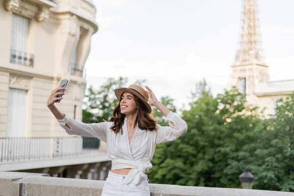 Positive traveler in sun hat taking selfie on smartphone with Eiffel tower at background in France — Stock Photo