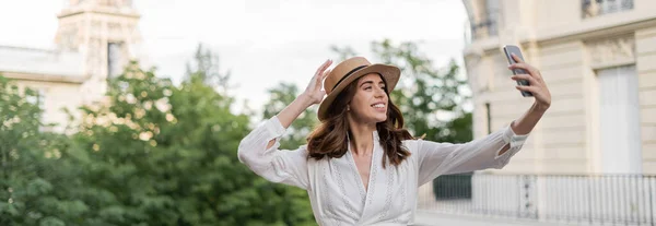 Весела молода жінка в сонцезахисному капелюсі робить селфі на смартфоні з розмитою вежею Ейфеля на задньому плані у Франції, банер — стокове фото