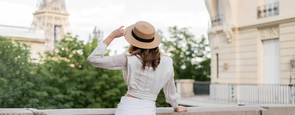 Back view of young brunette woman holding sun hat while looking at Eiffel tower in Paris, banner — Stock Photo