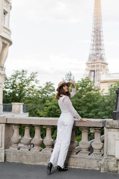 Glückliche junge Frau mit Sonnenhut blickt in die Kamera mit Eiffelturm im Hintergrund in Paris — Stockfoto