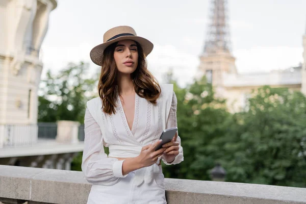 Elegante mujer con sombrero de sol sosteniendo el teléfono inteligente y mirando a la cámara con la torre Eiffel en el fondo en París - foto de stock