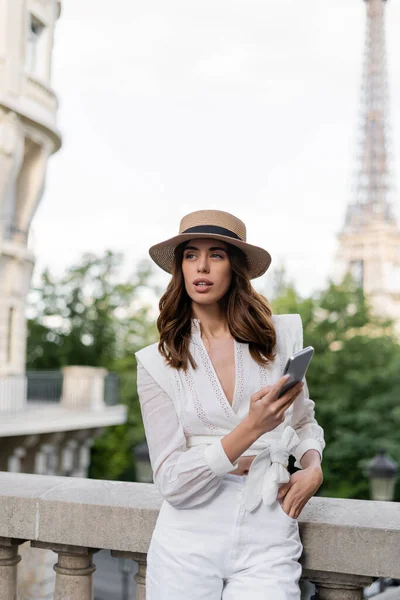 Femme branchée en chapeau de soleil tenant smartphone avec tour Eiffel à l'arrière-plan à Paris — Photo de stock