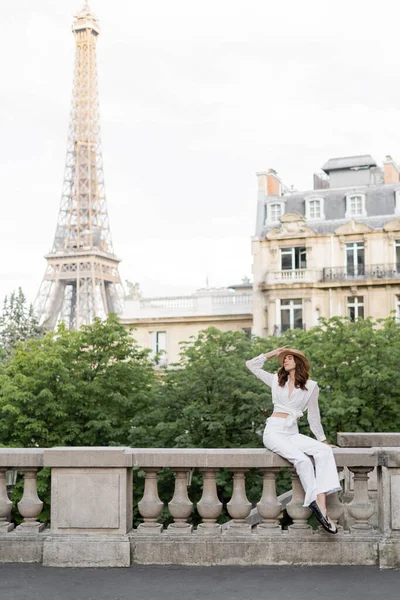 Elegante mujer sosteniendo sombrero de sol en la calle con la torre Eiffel al fondo en París - foto de stock