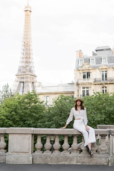 Mujer de moda sosteniendo teléfono inteligente y mirando a la cámara en la calle con la torre Eiffel en el fondo en París - foto de stock